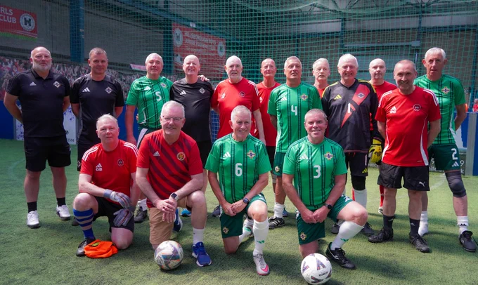 Health Minister Mike Nesbitt with a group of people in football kits. Some are kneeling, others are standing on grass. The football shirts are different colours including green, red and black.