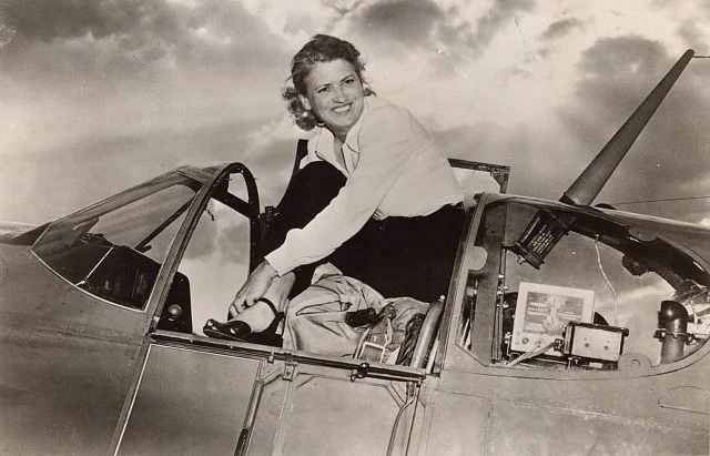 Woman sitting on the cockpit of a vintage aircraft, smiling at the camera under a cloudy sky.
