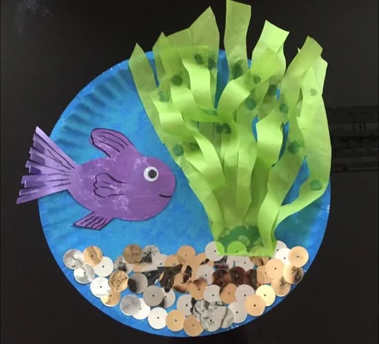 A Paper Plate Ocean Craft with a blue plate, green tissue paper seaweed, sparkly sequin sand, and a colorful fish, displayed on a table.