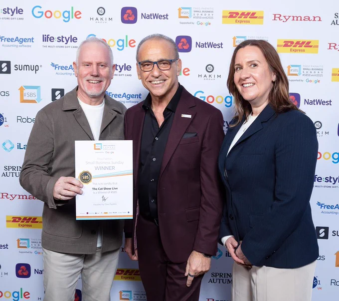 Three individuals posing with a "Small Business Sunday" award at an event sponsored by DHL, NatWest, and other brands. The central figure is Theo Paphitis, flanked by two representatives, one holding the award plaque.