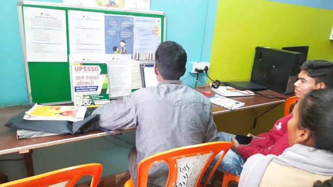 Young students at Bansa Library engaged in focused study around computers and books, with a librarian guiding them. The setting is calm and welcoming, reflecting a community space where local youth access resources, mentorship, and a supportive environment for learning and exam preparation without leaving their village.