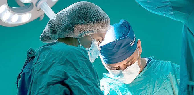 Two surgeons wearing gowns and masks are looking down as though operating on a patient.