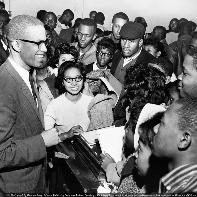 A black-and-white of Malcolm X is he signs an autograph for a young people in a crowded room.
