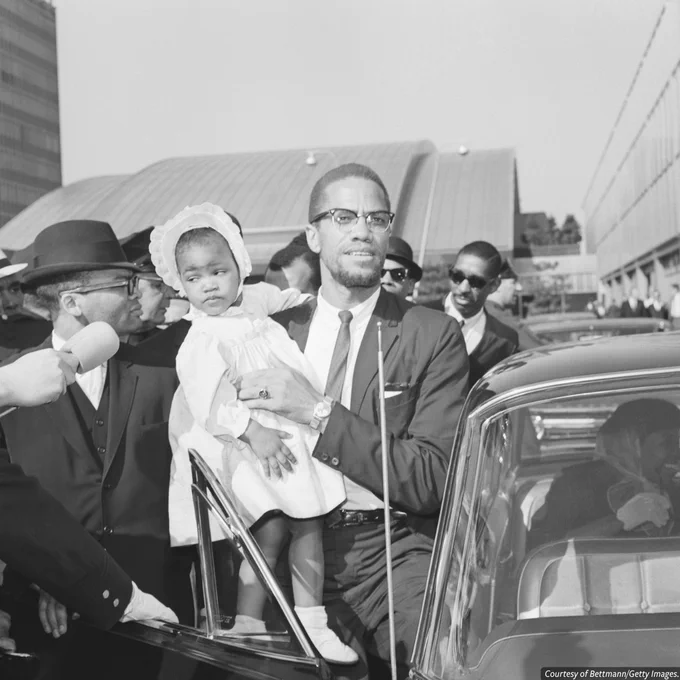A black-and-white photo of Malcolm X holding his child, standing beside a car in a crowded area.