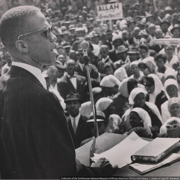 A black-and-white photo of Malcolm X as he speaks into a microphone at a public event, with a crowd listening in the background and a sign that reads “Allah is God” visible.