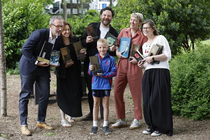 5 adult and one child stand in a clearing with lots of greenery. They are all holding a mixtures of trophys and books and smiling at the camera