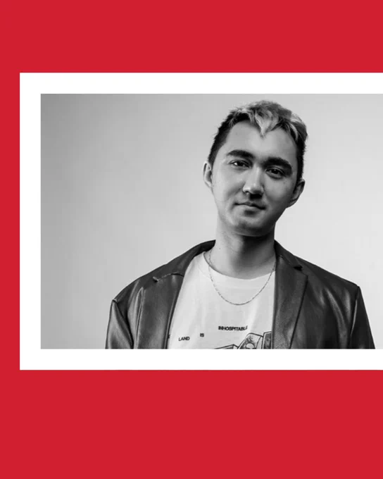 Black-and-white photo of Rob Macaisa Colgate, a multiracial person with short blond and black hair. They are wearing a leather jacket and a graphic T and standing with their thumbs in pant pockets. Photo is inset with a white border and red background beyond that.