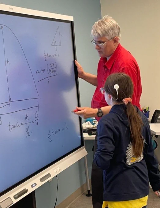 a man in a red shirt draws on a whiteboard with a girl standing with him and watching him. 