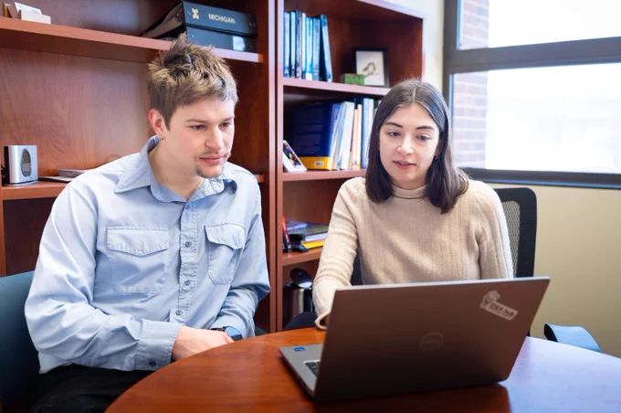 Left to right, Nathan Price and Anna Stuhlmacher are seated side by side at a table, looking at a laptop computer screen. 