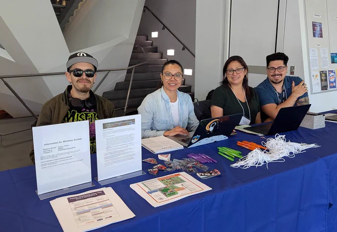 This is a group photo of four people sitting behind the registration table at the PRIMA working meeting, held in the Cahill Center for Astronomy and Astrophysics on the Caltech campus. The table holds brochures, lanyards, and swag (pens, stickers, magnets, etc.) for event attendees. From left to right: Daniel "Pina" Muro, Teresa Molano, Nancy Solis, and Sean Kindt. Image credit: Marcy Harbut, Caltech/IPAC