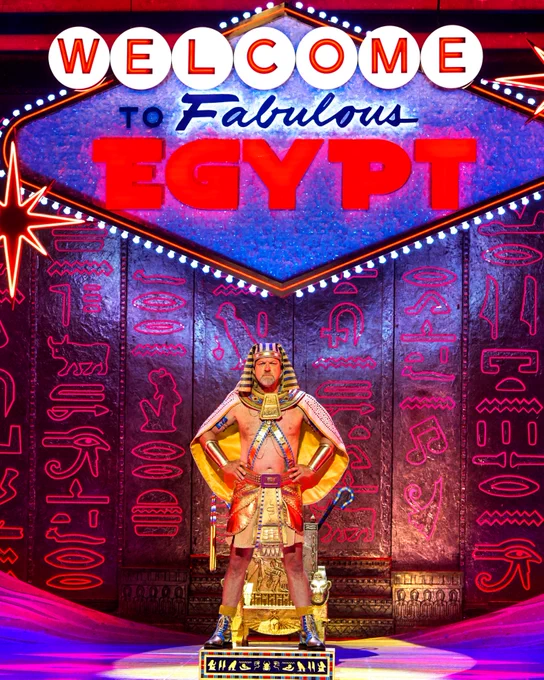 A person dressed in an elaborate Egyptian pharaoh costume stands in front of a vibrant, neon-lit backdrop that reads "Welcome to Fabulous Egypt," styled similarly to the iconic "Welcome to Fabulous Las Vegas" sign. The backdrop features hieroglyphics and traditional Egyptian symbols.