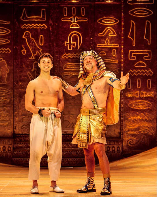 Two actors on stage in a scene from the musical "Joseph and the Amazing Technicolor Dreamcoat," with one dressed as an ancient Egyptian ruler and the other wearing a simple costume, both smiling and posing under warm stage lighting with hieroglyphic decorations in the background.
