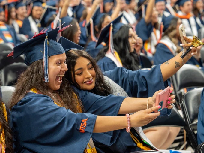 Two graduates in blue caps and gowns taking a selfie during the University of Texas at San Antonio commencement ceremony.