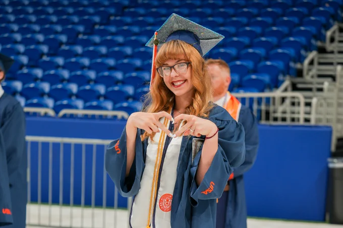 Graduate in a blue cap and gown joyfully displaying a diploma in a stadium setting.
