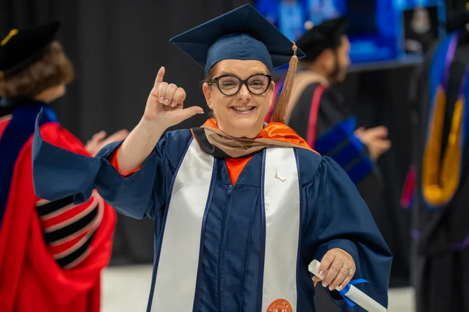 Graduate in blue and orange regalia giving a birds up at a graduation ceremony.