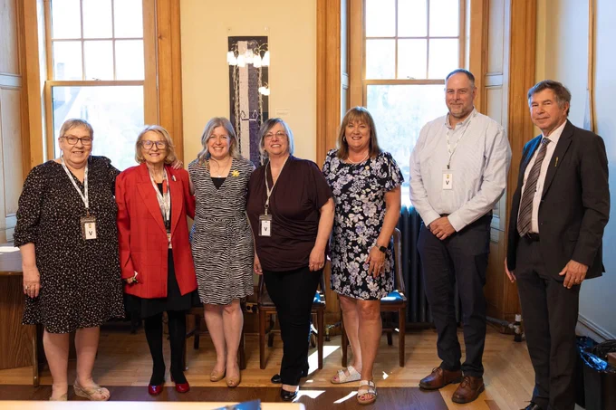 Seven adults stand together smiling in a well-lit room with large windows and wooden trim, wearing business or semi-formal attire and conference badges.