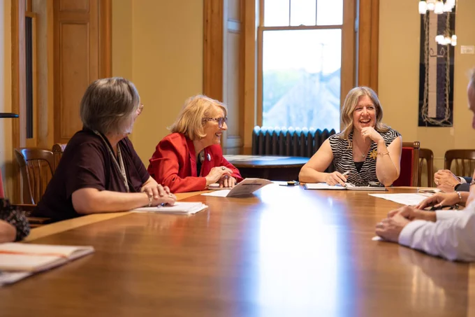 Three women sit at a conference table, engaged in conversation and smiling, with papers and pens in front of them. Sunlight comes through a window in the background, creating a warm, friendly atmosphere.