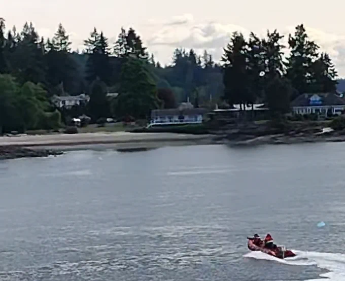 A rescue boat with people in red life vests on a coastal waterway, with houses and trees in the background.