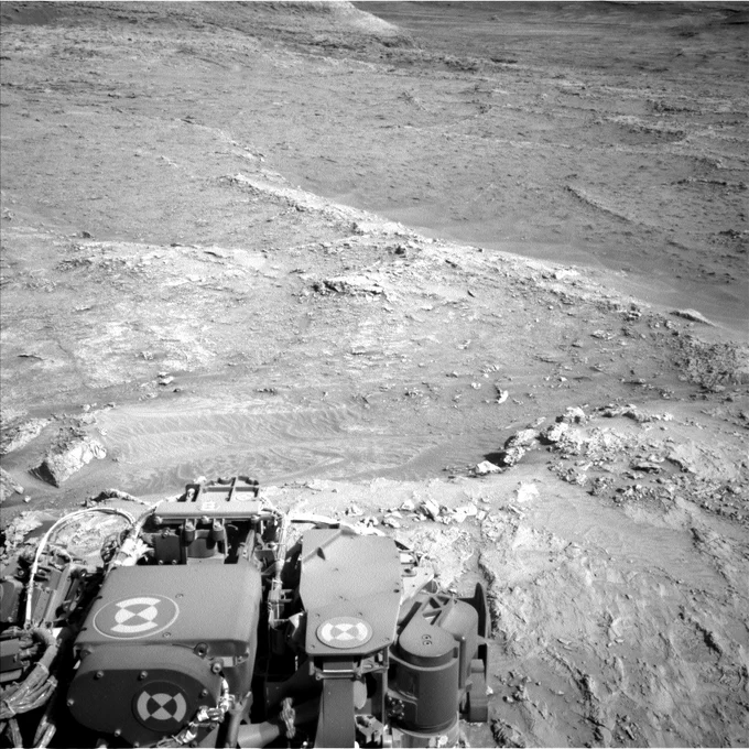 A black-and-white image taken by the Curiosity rover shows a rocky, uneven Martian landscape in Gale Crater. The rover's instrument deck is visible at the bottom of the frame. In the middle distance, rippled sand textures stretch across a small patch of terrain, with scattered rocks and ledges leading up to a gentle slope in the background. The scene is lit by soft sunlight, casting faint shadows across the surface.