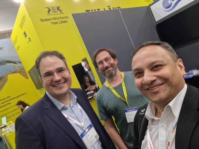 Drei Männer auf der Messe The Smarter E 2025 in München vor dem gelben Gemeinschaftsstand von „THE LÄND“ (Baden-Württemberg). Sie lächeln in die Kamera, tragen Namensschilder und stehen im Austausch am Stand.