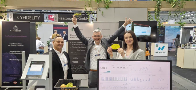Gruppenfoto an einem Messestand auf der The Smarter E 2025: Zwei Männer und eine Frau stehen vor digitalen Displays und einem Bildschirm mit Energieverbrauchsgrafiken. Eine Person hält ein Schild mit „HALLO“, eine andere mit „Nett hier“. Im Hintergrund Banner von CYFIDELITY und reasonance GmbH sowie weitere Messebesucher.