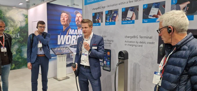 Ein Mitarbeiter von chargeBIG erklärt auf der Messe The smarter E 2025 in München die Funktionsweise einer Ladelösung für Elektrofahrzeuge. Er hält ein Ladekabel in der Hand und spricht ins Mikrofon. Um ihn herum stehen mehrere Messebesucher mit Headsets und hören aufmerksam zu. Im Hintergrund ist eine Anleitung zur Nutzung der chargeBIG App sowie ein Werbeplakat zu sehen.