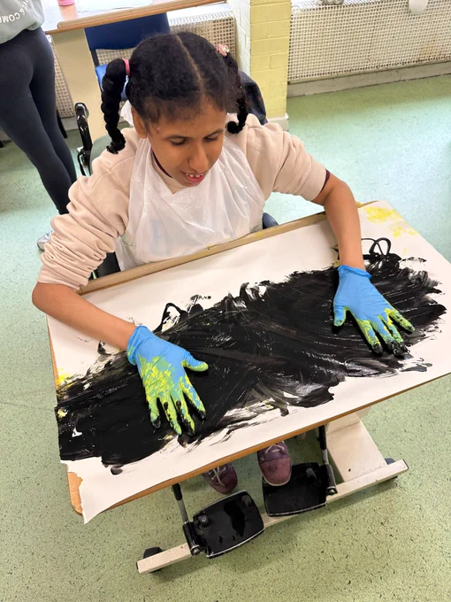 A young woman wearing blue gloves at at a table painting with her hands using black paint. 