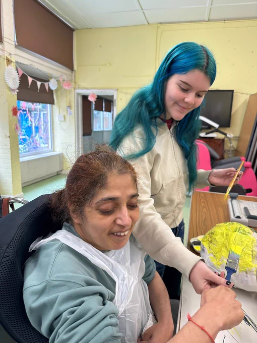 A young woman sat at a table with a paintbrush painting a papier mache ball in yellow, and a Support Worker stood beside her helping her hold the brush. 