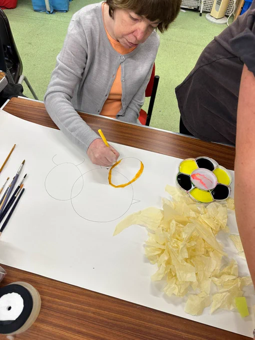 A woman sat at a table colouring in a bee shape with a yellow crayon.