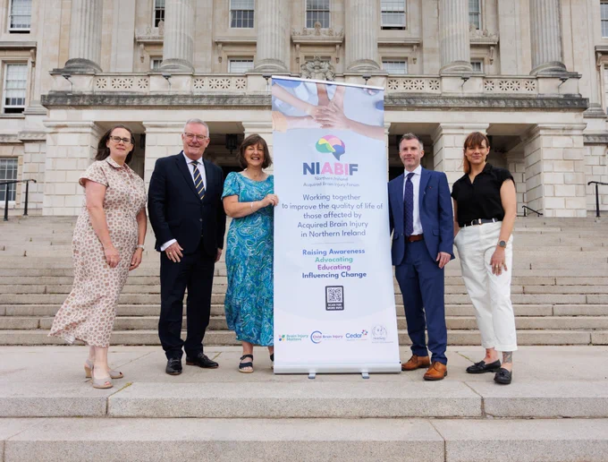 Elaine ArmstronG Cedar CEO, Health Minister Mike Nesbitt, Melanie Bowden Headway, Gerard Anderson Chair NIABIF, Dr Sharon Buckland Child Brain Injury Trust on the steps on Parliament Buildings with a NIABIF Stand. All are smiling at camera. It is a bright day.