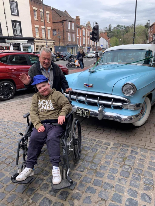 A man in a wheelchair with a man standing behind him, posing in front of an old fashioned blue car on a cobbled street. 