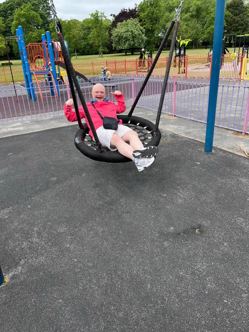 A man in a bright pink coat and white shorts swinging on a swing at a park. He's leaning back and giving a big smile and double thumbs up to the camera. 