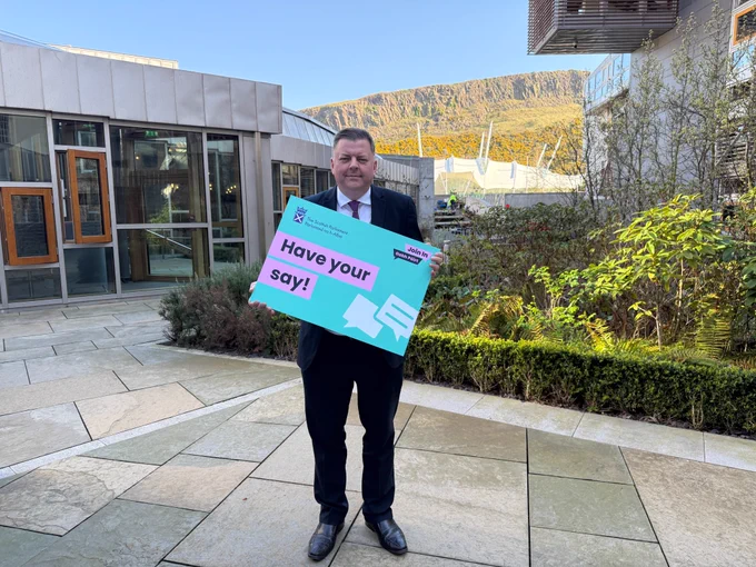 Colin Smyth MSP,  Convener of the Economy and Fair Work Committee, is  in the members'  garden of the Scottish Parliament. He is holding a brightly coloured sign that says 'Have your say!'