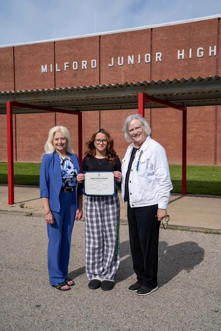 Student holding certificate in front of MJHS next to woman in blue and woman in white
