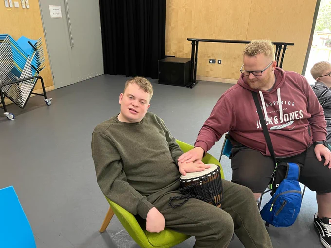 Two men are sitting next to each other on chairs. One is wearing a green sweatshirt and is holding a bongo drum in his lap. The man next to him wearing a burgundy sweatshirt is holding the other mans hand on the drum