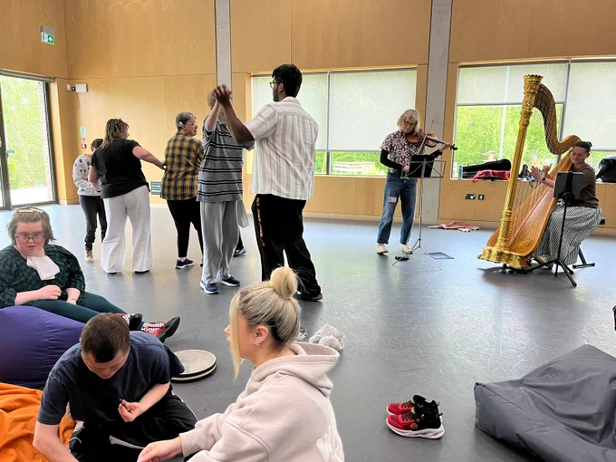 A group of people taking part in a music session. On the left there are five people standing up, dancing. On the right, a woman plays the violin and another plays the harp. In the foreground, three people sit on bean bags on the floor.  