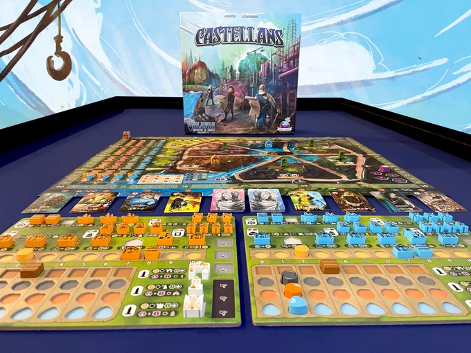 The box for the board game "Castellans" is in the back. In front is the game set up with a game board and two player boards that are adorned with many tokens representing buildings and ships.