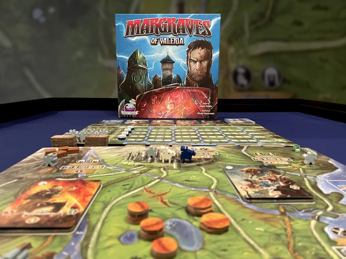 the box for the board game Margraves of Valeria is in the back and in front the board set up. Tokens, cards and margrave tokens are placed atop the board.