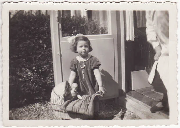 A vintage black-and-white photo showing a young child standing outside beside an open door. The child is wearing a light-colored dress with a dark ribbon and appears to be holding something small in one hand. On the ground next to the child is a small pushing chair with a doll. The edge of the photo is scalloped. Part of an adult, presumably walking out of the frame, is visible on the right side of the image.
