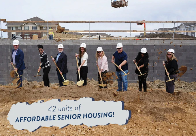 Eight people in hard hats and formal attire throwing dirt with shovels at a groundbreaking ceremony, with construction equipment and partially built homes in the background. Graphical text on top reads, "42 units of affordable senior housing!"