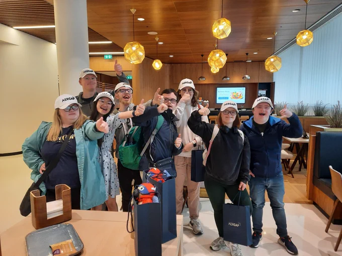 A group of TCPID students all wearing white caps with AerCap written on them. All are giving the thumbs up sign. They are also holding navy AerCaps bags with treats in them. 