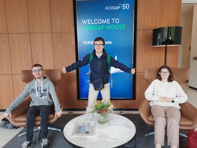 Two TCPID students are sitting in brown leather chairs. Another student is standing between the chairs with his arms out giving the thumbs up sign. Behind them a sign saying Welcome to AerCap House TCPID students is visible. 