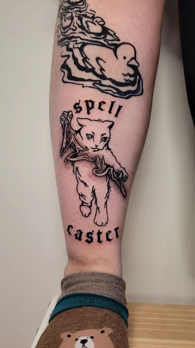  fresh tattoo on the front lower shin of a person. it is a cat wizard is casting lightening magic between its paws. text is arched above and below that reads, "spell caster". it is render in a blackwork style with a devious expression and the magic lightening is created with a stippling effect.
