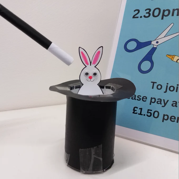 Photo of an upside-down black top hat made from black card with a white rabbit with googly eyes and pink and black drawn on detail sticking out of the hat. A classic magician's black and white wand made from rolled-up paper is sticking into the picture from the left, and to the right is part of a craft poster with black text on blue background and with graphics of scissors and crayons. The parts of the text that show read "2.30pm" and "£1.50 per child".