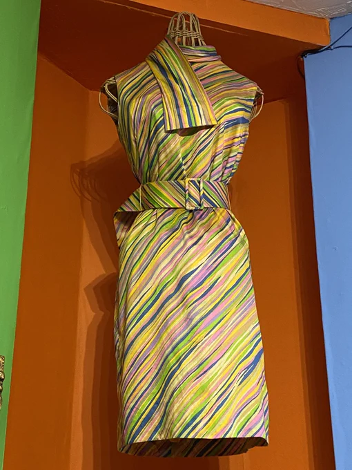 A woman’s 1969’s vintage shift dress, vibrant diagonal rainbow coloured fabric. Elegantly cinched in at the waist with a fabric matching belt. The walls behind the dress exhibit the colour palette, painted in bright green, orange and sky blue.