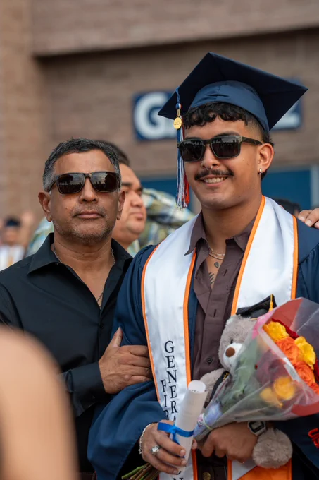 Graduate in cap and gown smiling next to an older adult at a graduation ceremony, holding a bouquet of flowers and a teddy bear, with a crowd in the background.