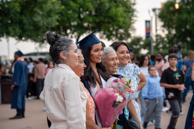 Graduate in cap and gown smiling with a bouquet of flowers, surrounded by three joyful family members at an outdoor graduation ceremony.