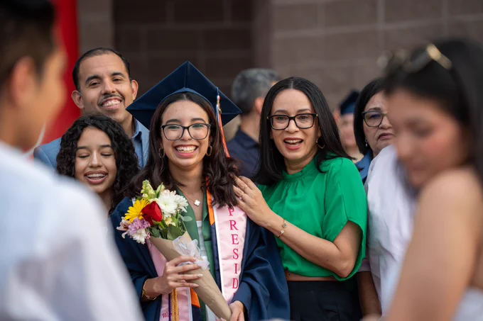 A joyful graduate holding flowers and wearing a cap and sash stands with a proud group of family members at a graduation ceremony.