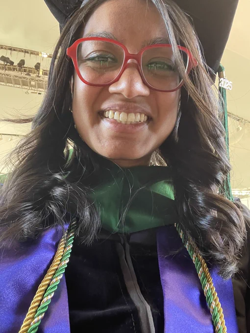 Closeup of Joya (brown skinned woman with shoulder length black hair and red glasses) smiling, wearing a black graduation gown and a green doctoral hood with a purple stole and green and gold cords around her shoulders