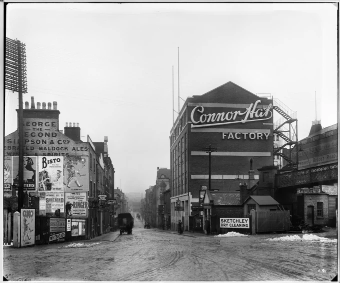 Black and white photo of a snowy street with a horse-drawn cart and the Connor Hats Factory building, surrounded by vintage shop signs and advertisements.
